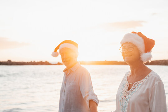 Old cute couple of mature persons enjoying and having fun together at the beach wearing christmas hats on holiday days. Walking on the beach with the sunset at the background at winter..