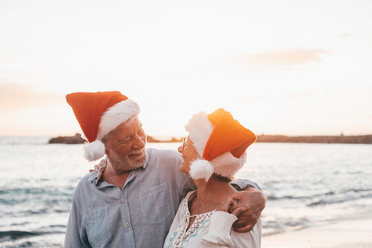 Old cute couple of mature persons enjoying and having fun together at the beach wearing christmas hats on holiday days. Hugged on the beach with the sunset at the background at winter..