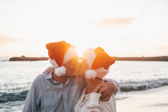 Old cute couple of mature persons enjoying and having fun together at the beach wearing christmas hats on holiday days. Hugged on the beach with the sunset at the background at winter..