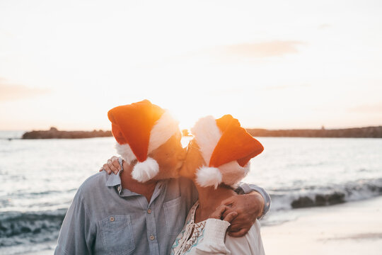 Old cute couple of mature persons enjoying and having fun together at the beach wearing christmas hats on holiday days. Hugged on the beach with the sunset at the background at winter.