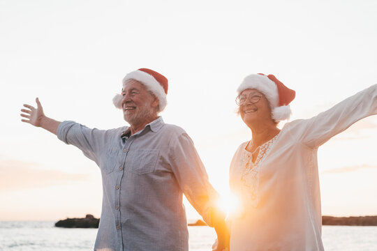 Old cute couple of mature persons enjoying and having fun together at the beach wearing christmas hats on holiday days. Walking on the beach with the sunset at the background at winter..