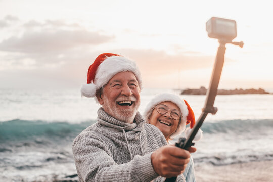 Portrait of two cute old persons having fun and enjoying together at the beach on christmas days at the beach wearing Christmas hats. Looking and holding a camera taking videos of vacations