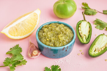 Bowl of tasty green salsa sauce and ingredients on pink background