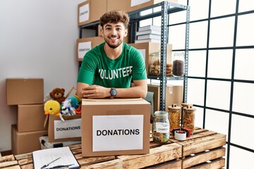 Young arab man wearing volunteer uniform leaning on donations cardboard box at charity center
