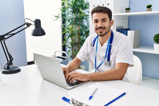 Young hispanic man wearing doctor uniform using laptop working at clinic