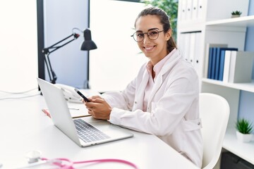 Young hispanic woman wearing doctor uniform using smartphone at clinic