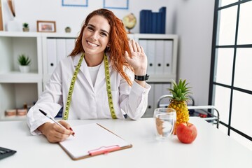 Young redhead woman nutritionist doctor at the clinic waiving saying hello happy and smiling,...