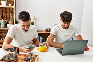 Fototapeta na wymiar Two hispanic men couple having breakfast using smartphone and laptop at home