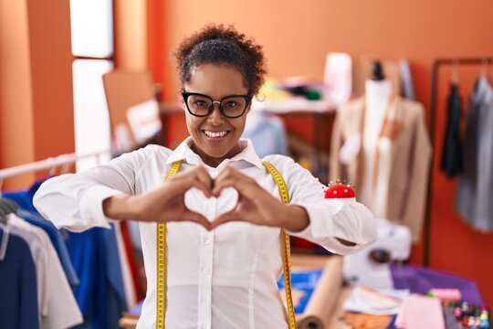 Young hispanic woman with curly hair standing at tailor room smiling in love showing heart symbol and shape with hands. romantic concept.