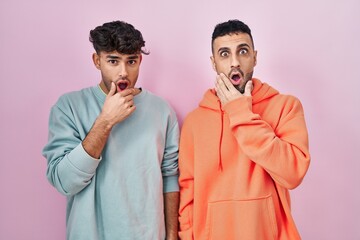 Young hispanic gay couple standing over pink background looking fascinated with disbelief, surprise and amazed expression with hands on chin