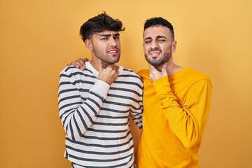 Young hispanic gay couple standing over yellow background touching painful neck, sore throat for flu, clod and infection