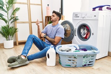 Young hispanic man putting dirty laundry into washing machine looking proud, smiling doing thumbs up gesture to the side