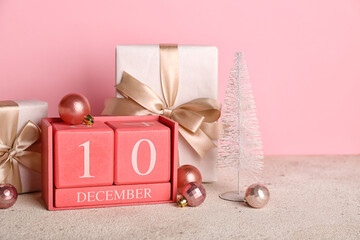 Cube calendar with date 10 DECEMBER, Christmas balls and gifts on table near pink wall