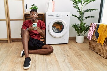 Young african american man using smartphone waiting for washing machine smiling looking to the side...