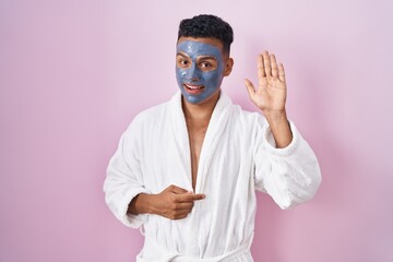 Young hispanic man wearing beauty face mask and bath robe waiving saying hello happy and smiling, friendly welcome gesture