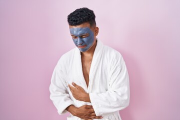 Young hispanic man wearing beauty face mask and bath robe with hand on stomach because indigestion, painful illness feeling unwell. ache concept.