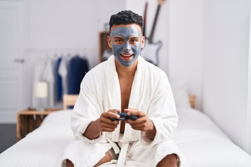 Young hispanic man wearing beauty face mask playing video games smiling and laughing hard out loud because funny crazy joke.