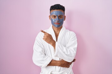 Young hispanic man wearing beauty face mask and bath robe pointing with hand finger to the side showing advertisement, serious and calm face