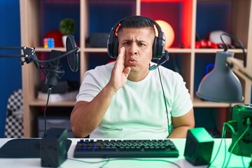 Young hispanic man playing video games hand on mouth telling secret rumor, whispering malicious talk conversation