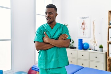 Young african physiotherapist man with crossed arms working at pain recovery clinic clueless and...