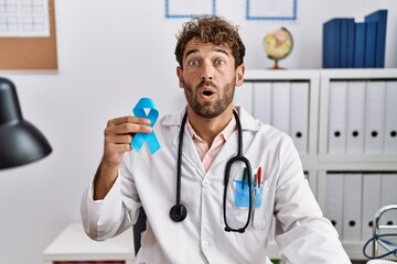 Young hispanic doctor man holding blue ribbon scared and amazed with open mouth for surprise, disbelief face