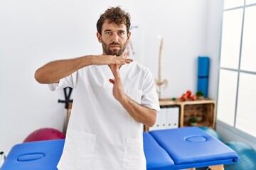 Young handsome physiotherapist man working at pain recovery clinic doing time out gesture with hands, frustrated and serious face