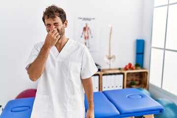 Young handsome physiotherapist man working at pain recovery clinic smelling something stinky and...