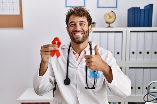 Young hispanic doctor man holding support red ribbon at medical clinic smiling happy and positive, thumb up doing excellent and approval sign