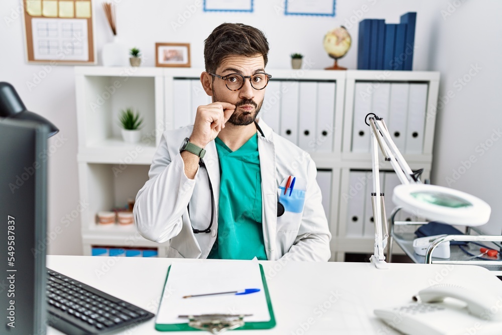 Sticker Young man with beard wearing doctor uniform and stethoscope at the clinic mouth and lips shut as zip with fingers. secret and silent, taboo talking