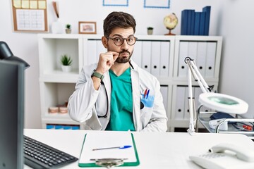 Young man with beard wearing doctor uniform and stethoscope at the clinic mouth and lips shut as...