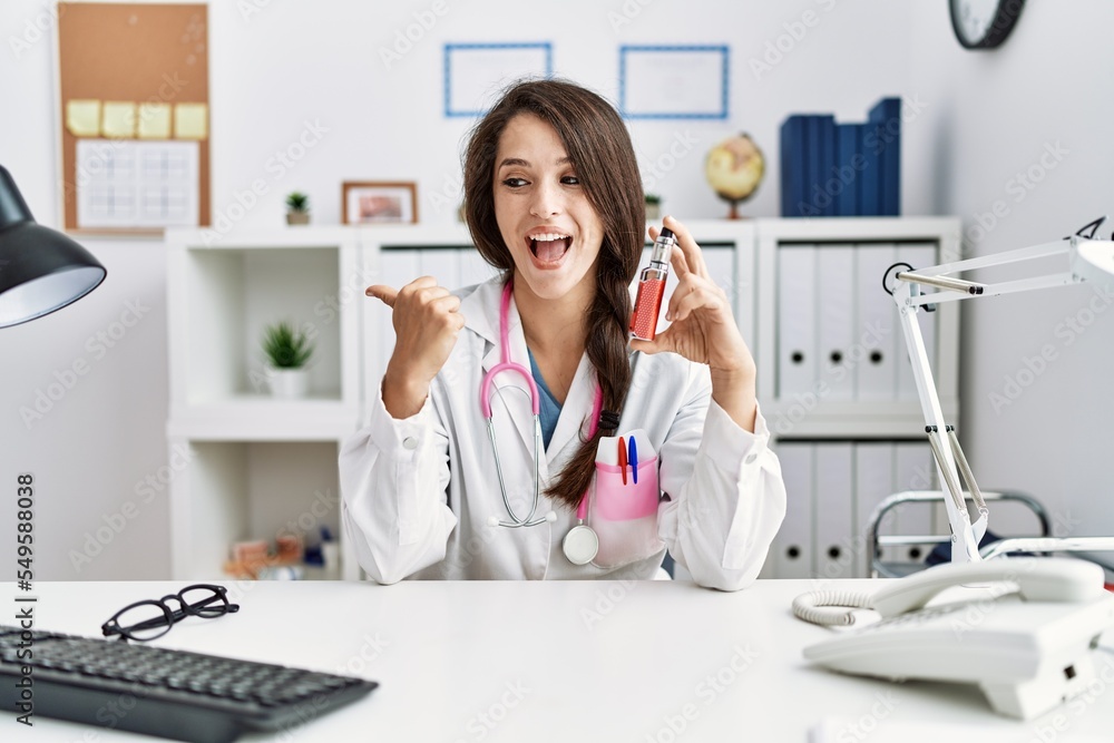 Sticker Young doctor woman holding electronic cigarette at medical clinic pointing thumb up to the side smiling happy with open mouth