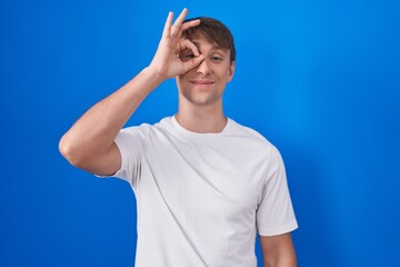 Naklejka premium Caucasian blond man standing over blue background doing ok gesture with hand smiling, eye looking through fingers with happy face.