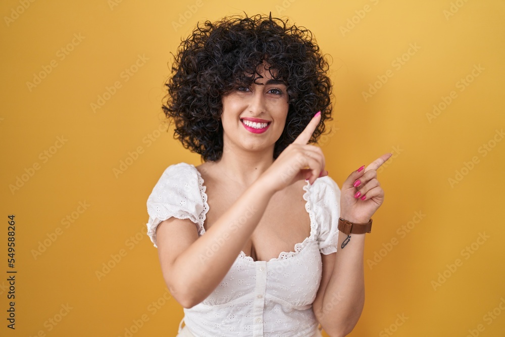 Poster young brunette woman with curly hair standing over yellow background smiling and looking at the came