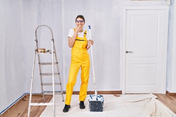 Young caucasian woman painting walls laughing and embarrassed giggle covering mouth with hands,...