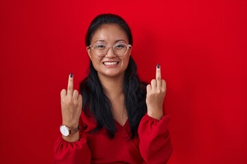 Asian young woman standing over red background showing middle finger doing fuck you bad expression,...