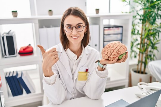 Young caucasian doctor woman holding brain as mental health concept smiling happy and positive, thumb up doing excellent and approval sign