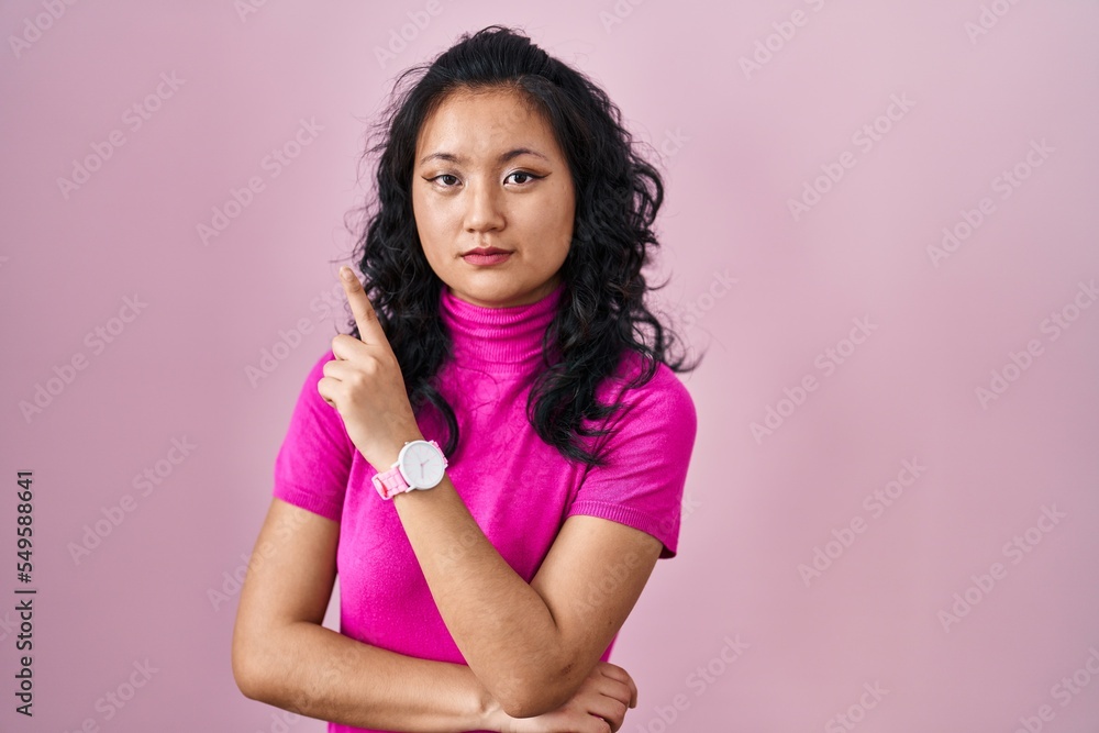 Sticker Young asian woman standing over pink background pointing with hand finger to the side showing advertisement, serious and calm face