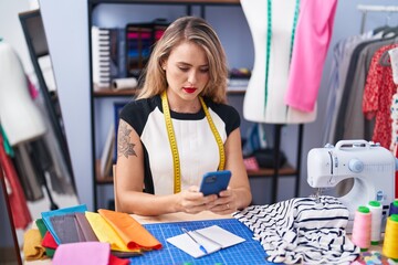 Young beautiful hispanic woman tailor using smartphone with serious expression at clothing factory