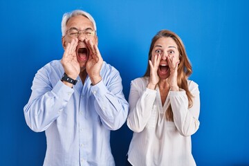 Middle age hispanic couple standing over blue background shouting angry out loud with hands over mouth