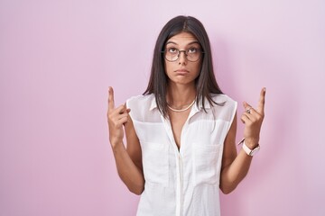 Brunette young woman standing over pink background wearing glasses pointing up looking sad and...