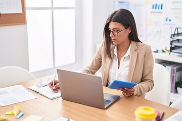 Young beautiful hispanic woman business worker using touchpad and laptop writing on notebook at office