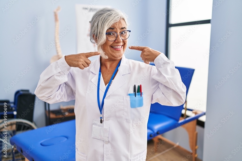 Poster Middle age woman with grey hair working at pain recovery clinic smiling cheerful showing and pointing with fingers teeth and mouth. dental health concept.