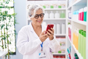 Middle age grey-haired woman pharmacist using smartphone working at pharmacy