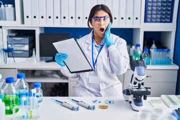 Fototapeta premium Hispanic young woman working at scientist laboratory afraid and shocked, surprise and amazed expression with hands on face
