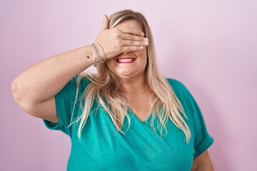 Caucasian plus size woman standing over pink background smiling and laughing with hand on face covering eyes for surprise. blind concept.