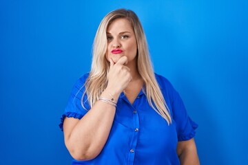 Caucasian plus size woman standing over blue background looking confident at the camera with smile with crossed arms and hand raised on chin. thinking positive.