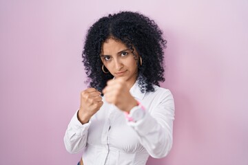 Hispanic woman with curly hair standing over pink background ready to fight with fist defense...