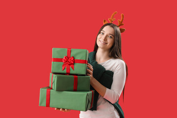 Young woman in reindeer horns with Christmas gifts on red background