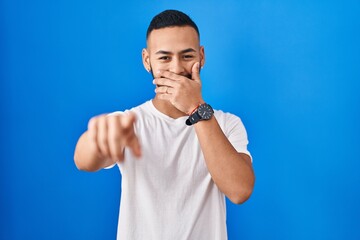 Young hispanic man standing over blue background laughing at you, pointing finger to the camera...