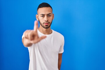 Naklejka premium Young hispanic man standing over blue background pointing with finger up and angry expression, showing no gesture